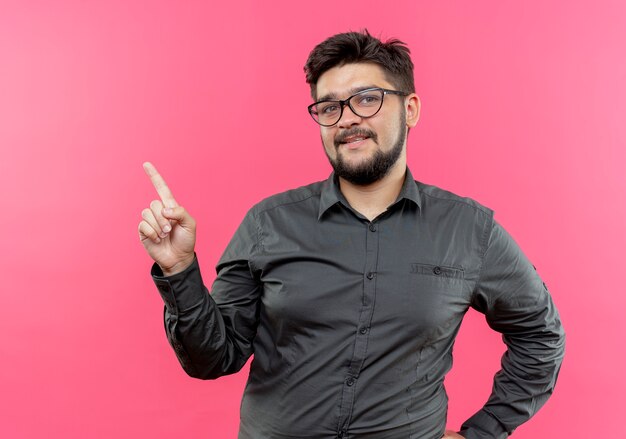 pleased young businessman wearing glasses points at side isolated on pink with copy space