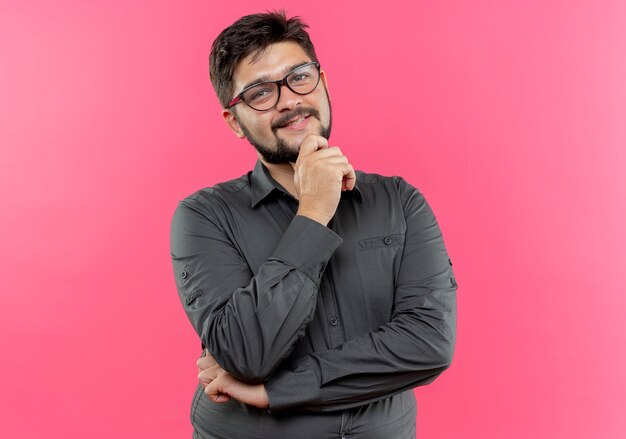 Pleased young businessman wearing glasses putting hand under chin isolated on pink wall