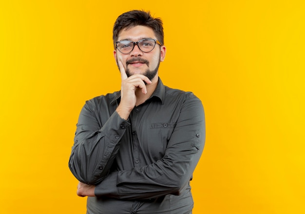 Pleased young businessman wearing glasses putting hand on chin isolated on yellow background