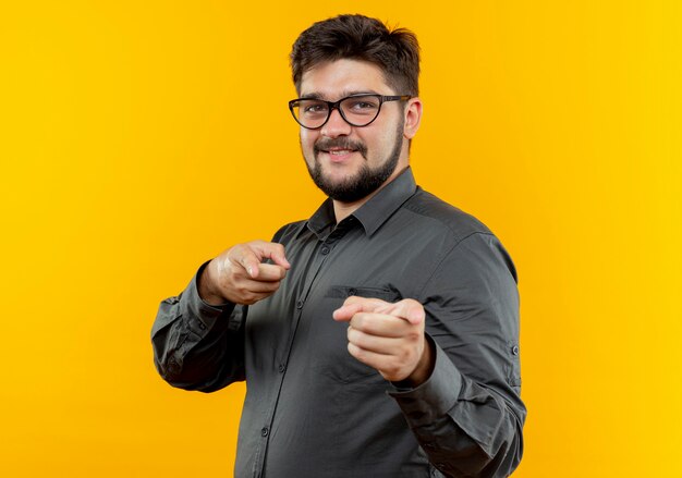 Pleased young businessman wearing glasses showing you gesture isolated on yellow background