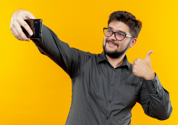 Pleased young businessman wearing glasses take a selfie his thumb up isolated on yellow background