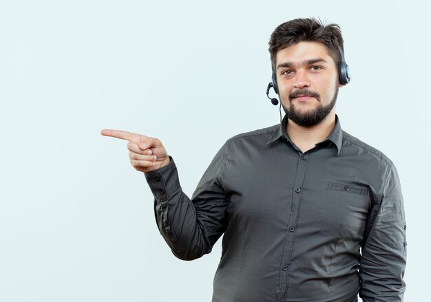 Pleased young call center man wearing headset points at side isolated on white background with copy space