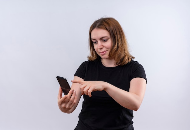 Pleased young casual woman holding mobile phone and pointing at it on isolated white space with copy space