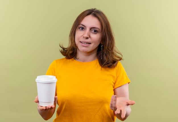 Pleased young casual woman holding plastic coffee cup and showing empty hand on isolated green space with copy space