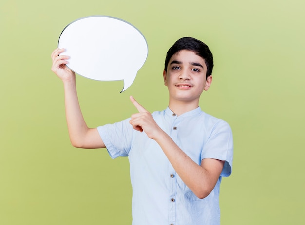 Pleased young caucasian boy holding and pointing at speech bubble looking at camera isolated on olive green background with copy space