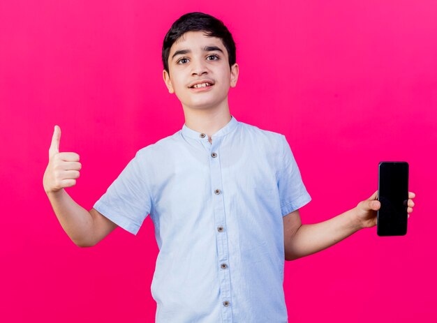 Pleased young caucasian boy showing mobile phone looking at camera showing thumb up isolated on crimson background