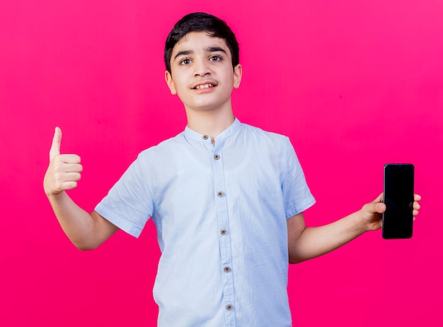 Довольный молодой кавказский мальчик показывает мобильный телефон, глядя в камеру, показывает палец вверх, изолированные на малиновом фоне