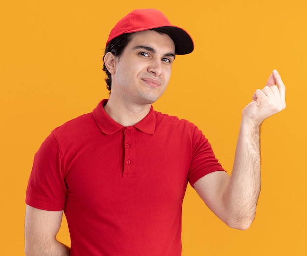 Felice giovane fattorino caucasico in uniforme blu e berretto che tiene la mano dietro la schiena facendo un gesto di punta isolato sul muro arancione