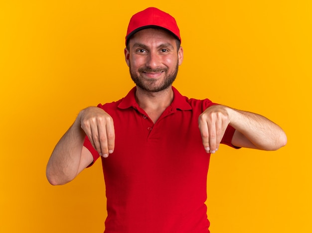 Foto gratuita felice giovane fattorino caucasico in uniforme rossa e berretto che guarda la telecamera fingere di tenere qualcosa di isolato su una parete arancione