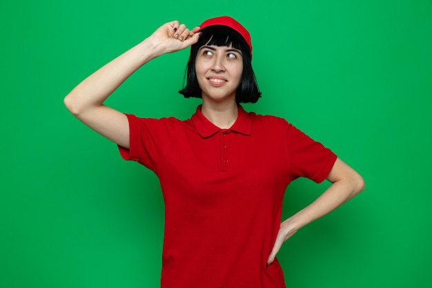 Free photo pleased young caucasian delivery woman holding her cap and looking at side