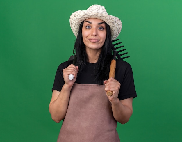 Pleased young caucasian gardener girl wearing uniform and hat holding rake and hoe-rake on shoulders 