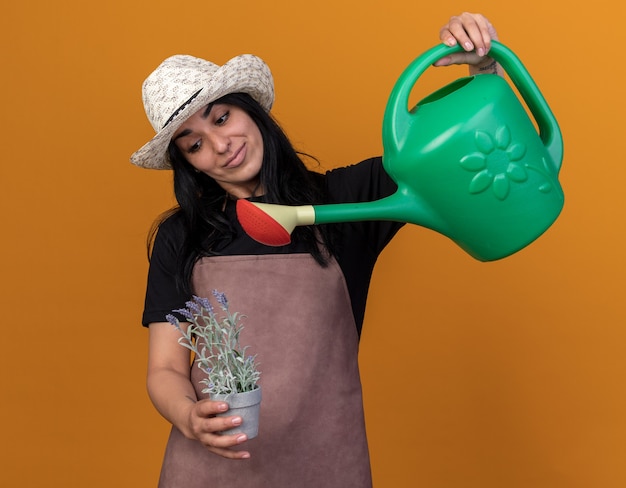 Pleased young caucasian gardener girl wearing uniform and hat watering flowers in `flowerpot with watering can 