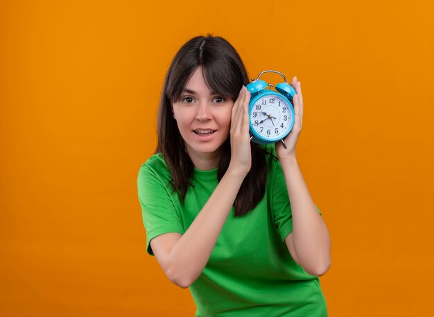 Pleased young caucasian girl in green shirt holds clock with both hands on isolated orange background