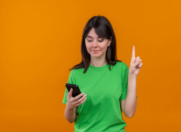 Pleased young caucasian girl in green shirt holds phone and points up on isolated orange background with copy space