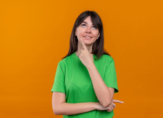 Pleased young caucasian girl in green shirt puts finger on chin and looks up on isolated orange background with copy space