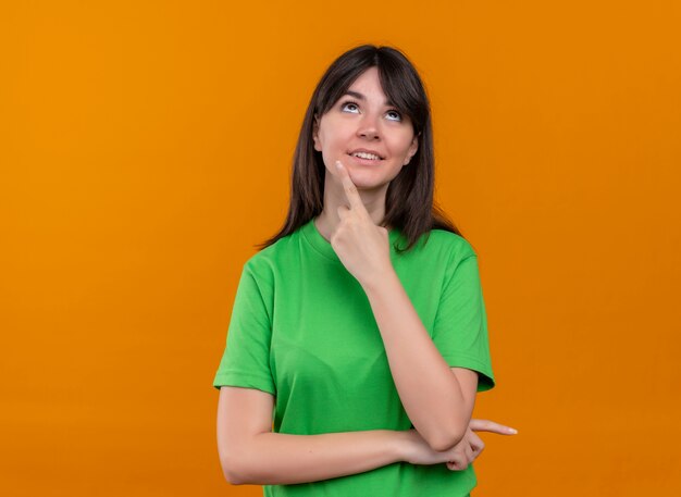 Довольная молодая кавказская девушка в зеленой рубашке кладет палец на подбородок и смотрит вверх на изолированный оранжевый фон с копией пространства