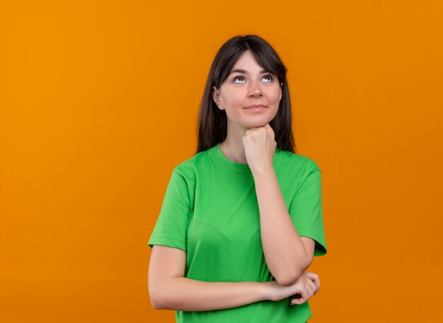 Pleased young caucasian girl in green shirt puts fist on chin and looks up on isolated orange background with copy space