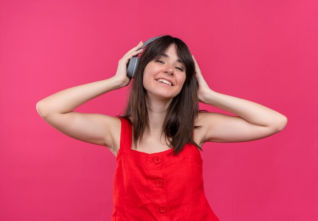 Pleased young caucasian girl holding headphones on head on isolated pink background