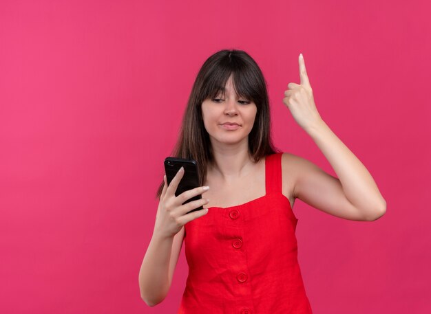 Pleased young caucasian girl holding phone and pointing up looking at phone on isolated pink background with copy space