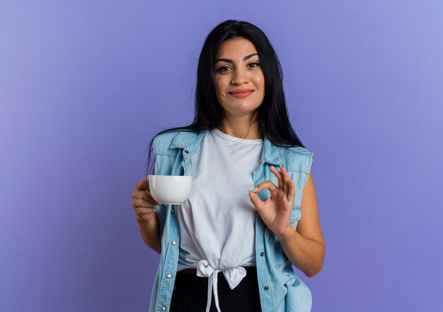 Pleased young caucasian girl holds cup and gestures ok hand sign isolated on purple background with copy space