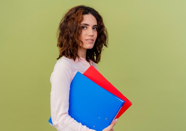 Pleased young caucasian girl stands sideways holding file folders isolated on green background with copy space