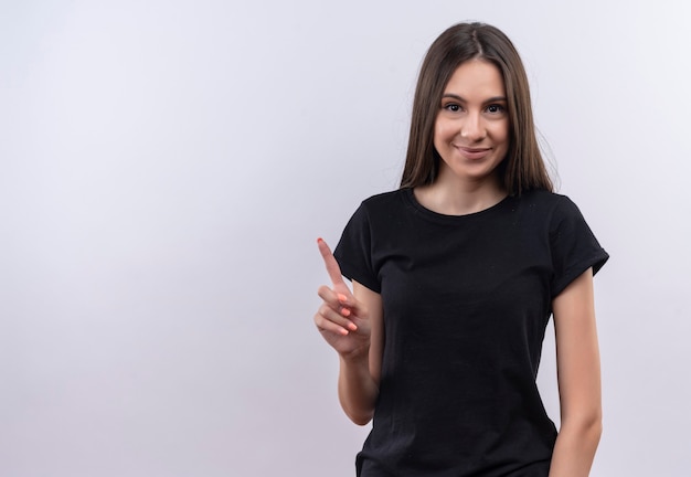 Довольная молодая кавказская девушка в черной футболке указывает пальцем на изолированную белую стену