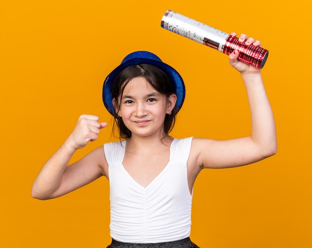 pleased young caucasian girl with blue party hat holding confetti cannon and keeping fist up isolated on orange wall with copy space