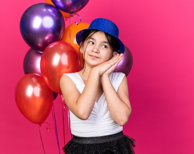 довольная молодая кавказская девушка в синей партийной шляпе, взявшись за руки и глядя на сторону, стоя перед гелиевыми шарами, изолированными на розовой стене с копией пространства