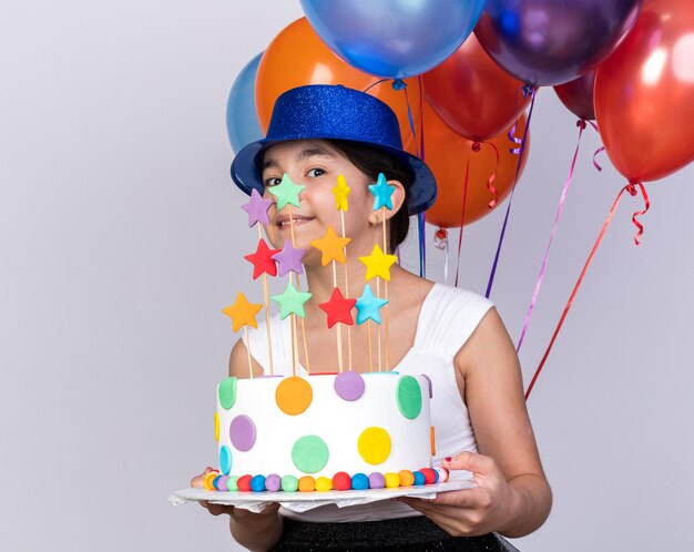 pleased young caucasian girl with blue party hat holding helium balloons and birthday cake isolated on white wall with copy space