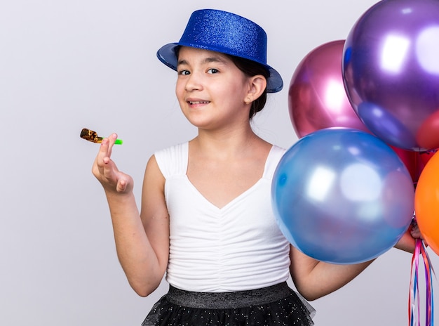 Contenta giovane ragazza caucasica con cappello da festa blu che tiene palloncini di elio e fischio di festa isolato sul muro bianco con spazio di copia