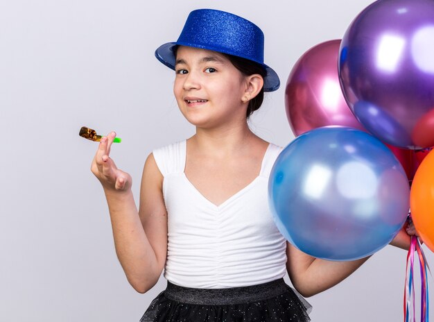 Довольная молодая кавказская девушка в синей партийной шляпе держит гелиевые шары и партийный свисток на белой стене с копией пространства