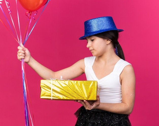 довольная молодая кавказская девушка в синей партийной шляпе смотрит на подарочную коробку и держит гелиевые шары, изолированные на розовой стене с копией пространства