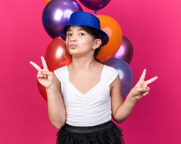 pleased young caucasian girl with blue party hat standing in front of helium balloons gesturing victory sign isolated on pink wall with copy space