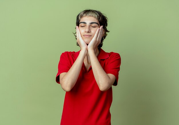 Pleased young caucasian girl with pixie haircut putting hands on face with closed eyes isolated on olive green background with copy space