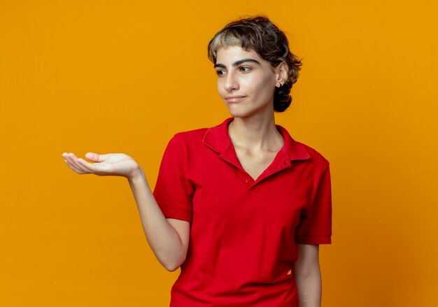 Pleased young caucasian girl with pixie haircut showing empty hand looking at side isolated on orange background with copy space