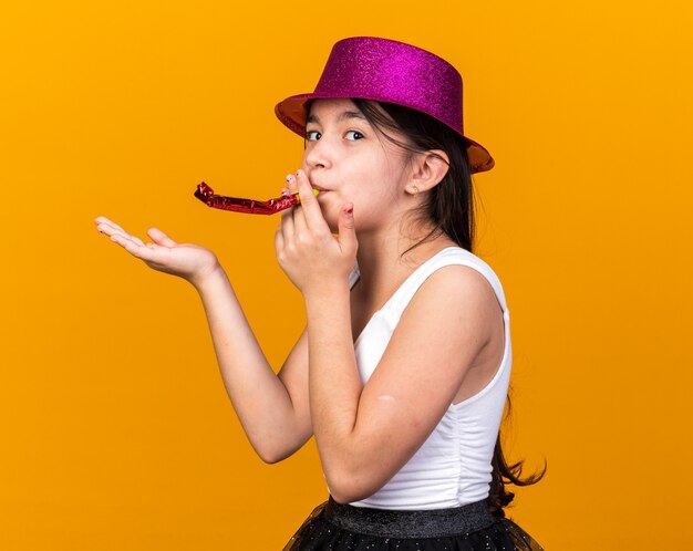 pleased young caucasian girl with purple party hat blowing party whistle and keeping hand open isolated on orange wall with copy space