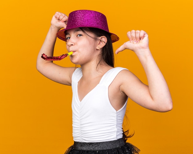 pleased young caucasian girl with purple party hat blowing party whistle and pointing at herself isolated on orange wall with copy space