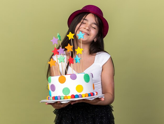 pleased young caucasian girl with purple party hat holding birthday cake isolated on olive green wall with copy space