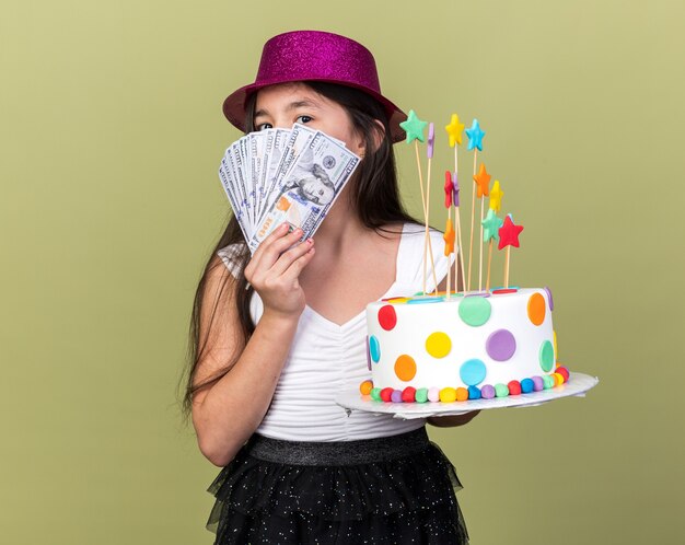 생일 케이크와 복사 공간 올리브 녹색 벽에 고립 된 돈을 들고 보라색 파티 모자와 함께 기쁘게 젊은 백인 여자