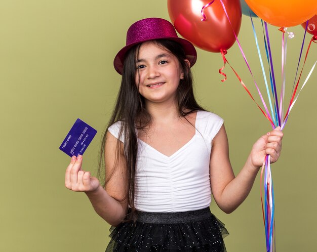 Contenta giovane ragazza caucasica con cappello da festa viola in possesso di carta di credito e palloncini di elio isolati su parete verde oliva con spazio di copia