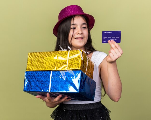 pleased young caucasian girl with purple party hat holding gift boxes and looking at credit card isolated on olive green wall with copy space
