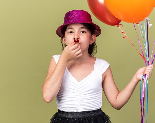 pleased young caucasian girl with purple party hat holding helium balloons and blowing party whistle isolated on olive green wall with copy space