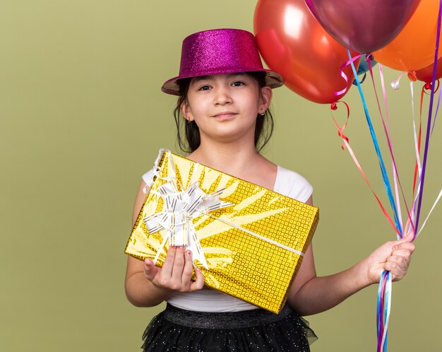 pleased young caucasian girl with purple party hat holding helium balloons and gift box isolated on olive green wall with copy space