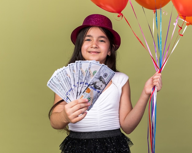 pleased young caucasian girl with purple party hat holding helium balloons and money isolated on olive green wall with copy space