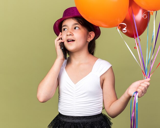 pleased young caucasian girl with purple party hat holding helium balloons talking on phone looking at side isolated on olive green wall with copy space