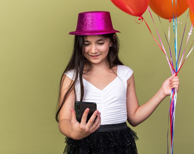 pleased young caucasian girl with purple party hat looking at phone and holding helium balloons isolated on olive green wall with copy space