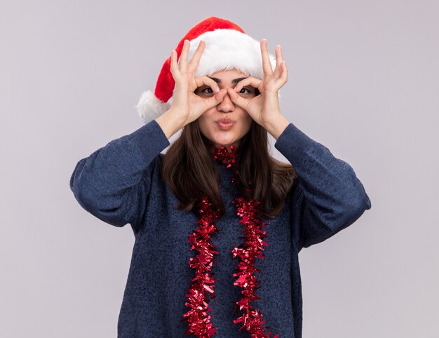 Pleased young caucasian girl with santa hat and garland around neck  through fingers isolated on white wall with copy space