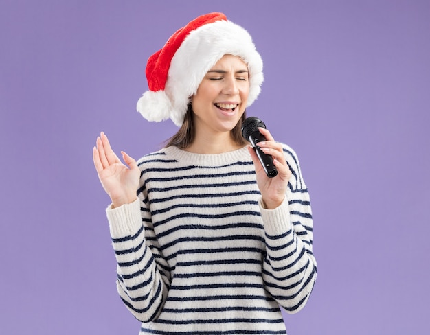 Free photo pleased young caucasian girl with santa hat holds mic pretending to sing isolated on purple background with copy space