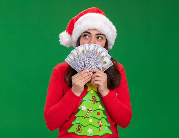 pleased young caucasian girl with santa hat holds money looking at side isolated on green background with copy space