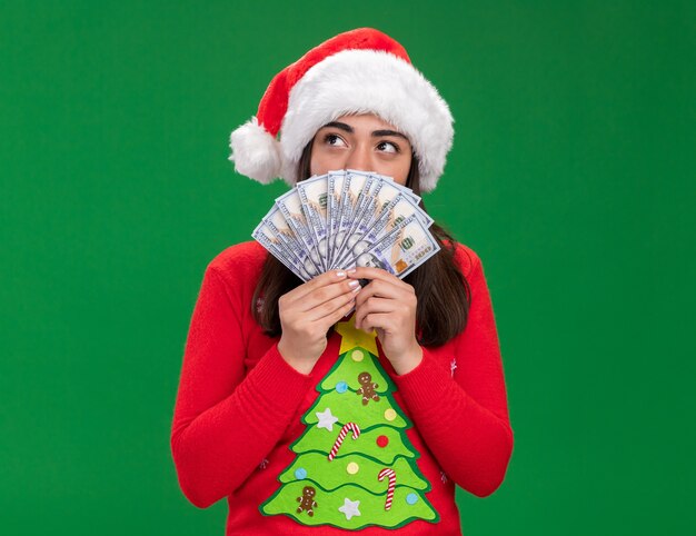 pleased young caucasian girl with santa hat holds money looking at side isolated on green background with copy space